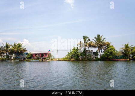 Alappuzha, Inde - 6 mars 2023 : plusieurs maisons riveraines aux extérieurs colorés et aux palmiers environnants sont visibles le long des backwaters sous un ciel clair. Banque D'Images