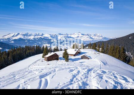 Chalets du Leutellet, France - 29 janvier 2023 : plusieurs chalets en bois sont vus sur une colline enneigée, entourée de pins, avec des chaînes de montagnes visibles en arrière-plan sous un ciel bleu clair. Banque D'Images