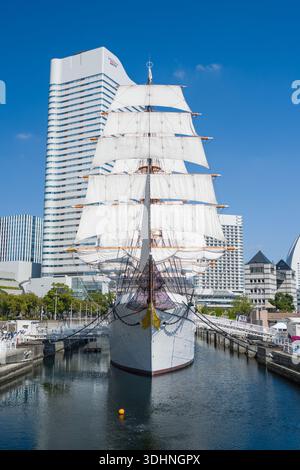Yokohama, Japon - 13 octobre 2024 : L'image montre une vue frontale du voilier commémoratif Nippon Maru amarré dans un canal, avec ses voiles blanches entièrement serrées, entouré de gratte-ciel modernes sous un ciel bleu clair. Banque D'Images