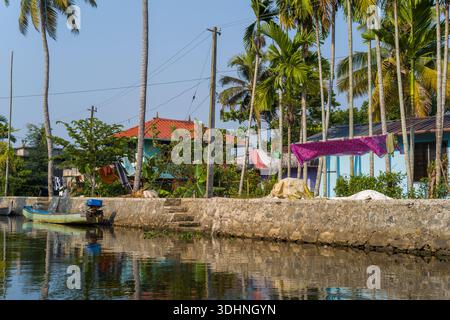 Alappuzha, Inde - 7 mars 2023 : le linge est suspendu pour sécher sur un remblai de pierre à côté d'un canal, avec des palmiers, des bananiers, des maisons résidentielles et de petits bateaux visibles sous un ciel dégagé. Banque D'Images