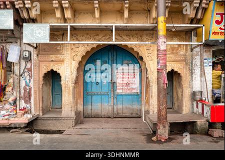 Mathura, Inde - 13 mars 2024 : la façade d'une maison historique présente des sculptures en pierre ornées, une grande double porte arquée bleue avec écriture hindi, flanquée de deux portes arquées plus petites, avec des magasins de rue adjacents et un homme visible à l'intérieur d'un magasin pendant la journée. Banque D'Images