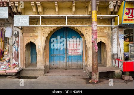 Mathura, Inde - 13 mars 2024 : la façade d'une maison historique présente des sculptures en pierre ornées, une grande double porte arquée bleue avec écriture hindi, flanquée de deux petites niches arquées, avec des magasins de rue adjacents, une signalisation commerciale, et un homme visible à l'intérieur d'un magasin pendant la journée. Banque D'Images