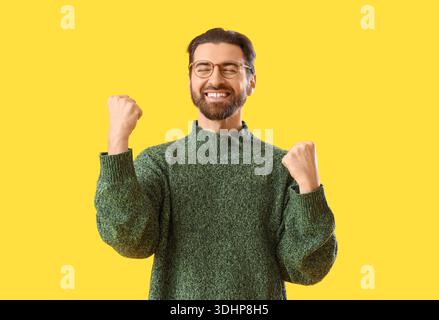 Portrait de jeune homme heureux en pull chaud célébrant le succès sur fond jaune Banque D'Images