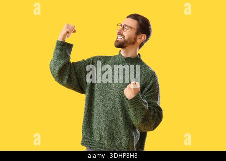 Portrait de jeune homme heureux en pull chaud célébrant le succès sur fond jaune Banque D'Images