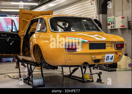 Vue arrière d'une Alfa Romeo 1750 GTAm 1971 orange-jaune classique dans un garage de course, surélevée sur des crics avec le capot et la porte ouverts, prêt pour l'entretien Banque D'Images