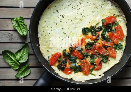 Une omelette salée ou frittata avec épinards frais et tomates cerises dans une poêle. Fond en bois. Vue de dessus. Banque D'Images