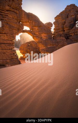 Vue de la lumière dorée perçant à travers une arche naturelle de formations rocheuses accidentées, jetant des ombres sur les dunes de sable ondulées, Djanet, province d'Illizi, Alg Banque D'Images