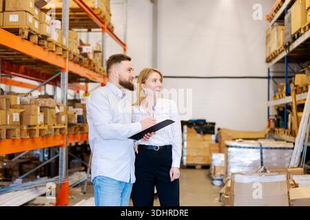 Hommes et femmes superviseurs logistiques debout dans un entrepôt, tenant une planchette à pince et discutant des niveaux de stock. Travail d'équipe professionnel dans un grand stockage Banque D'Images