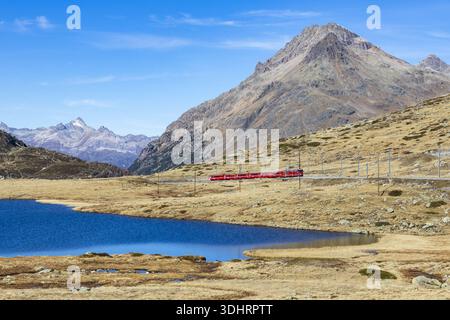 Vue d'un train rouge vibrant coupant à travers le paysage serein, juxtaposé contre un lac bleu et des montagnes majestueuses, Passo del Bernina, Grisons, S. Banque D'Images