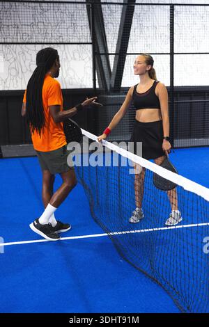 Homme afro-américain et femme asiatique serrant la main et saisissant des raquettes au filet sur le court de padel. Athlétique, travail d'équipe, dynamique, urbain, compétitif, fitness, communauté || autorisation du modèle Banque D'Images