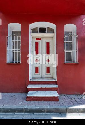Façade rouge vif avec une porte en bois blanc ornée de panneaux rouges, flanquée de deux fenêtres cintrées avec des barres décoratives en fer blanc et des marches Banque D'Images
