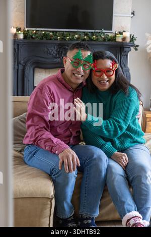 Souriant couple senior diversifié portant arbre de vacances et verres de bois assis sur le canapé près de la cheminée. Festif, confortable, célébration, chaleur, intimité, traditionnel, joyeux || modèle libéré Banque D'Images