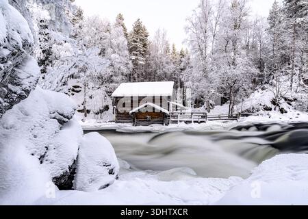 Vue sur une cabane rustique en bois nichée au milieu d'arbres enneigés le long d'une rivière ruisselante, une scène hivernale merveilleuse, Myllykoski, Finlande. Banque D'Images