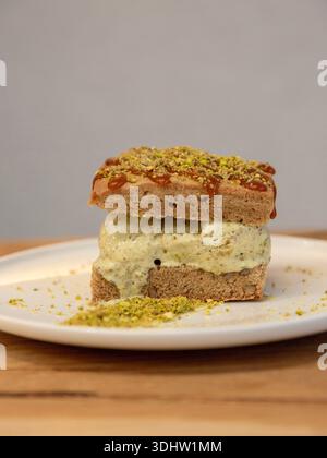 Tranche de dessert en couches de crème glacée à la pistache surmontée de pistache écrasée sur une assiette blanche, photographie de dessert sur une table en bois, vue de face. Banque D'Images