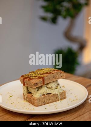 Tranche de dessert en couches de crème glacée à la pistache surmontée de pistache écrasée sur une assiette blanche, photographie de dessert sur une table en bois, vue de face. Banque D'Images