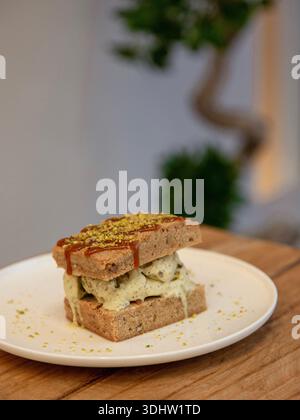 Tranche de dessert en couches de crème glacée à la pistache surmontée de pistache écrasée sur une assiette blanche, photographie de dessert sur une table en bois, vue de face. Banque D'Images