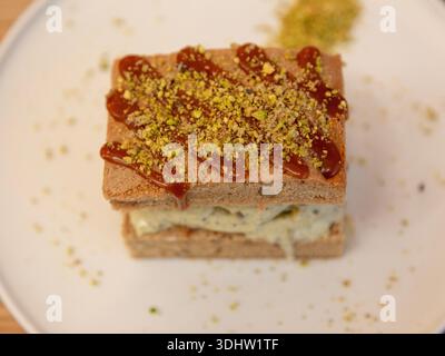 Tranche de dessert en couches de crème glacée à la pistache surmontée de pistache écrasée sur une assiette blanche, photographie de dessert sur une table en bois, vue de face. Banque D'Images