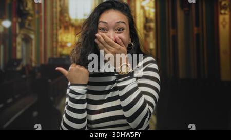 Jeune femme hispanique couvre la bouche avec la main et pointe le pouce à côté à l'intérieur de l'église ornée ; surprise amusée. Banque D'Images