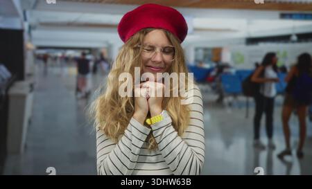 Femme se reposant les mains sous le menton et souriant à l'aéroport, jeune modèle blond ajustant les lunettes et béret rouge sur le pull rayé. Banque D'Images
