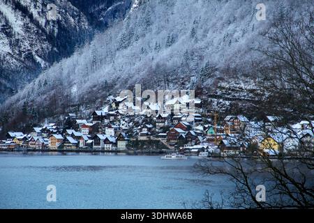Panorama hivernal du village de Hallstatt en Autriche, avec des maisons enneigées et des montagnes alpines reflétant dans les eaux calmes du lac à midi. Banque D'Images