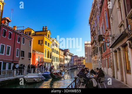 Touristes se relaxant dans un café en plein air à côté d'un canal pittoresque avec des bâtiments historiques colorés et des bateaux amarrés à Venise, en Italie, pendant l'hiver. Banque D'Images