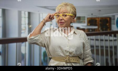 Femme hispanique senior touche des lunettes jaunes et pointe le doigt vers le temple dans le bâtiment ; sagesse réfléchie. Banque D'Images