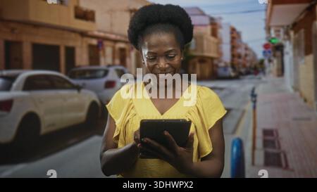 Femme tapant l'écran de la tablette avec le doigt sur le trottoir de la rue près des voitures garées, portant une robe jaune ; contentement focus. Banque D'Images