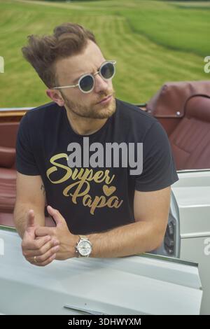 Homme portant des lunettes de soleil penché décontracté sur un ensemble convertible contre un champ vert pittoresque, portant le texte de super papa imprimé sur un T-shirt comme symbole de fierté Banque D'Images