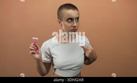 Femme aux cheveux courts tenant un rasoir jetable rose, pointe le doigt sur la poitrine tout en portant un haut blanc recadré en studio ; autonomisation. Banque D'Images