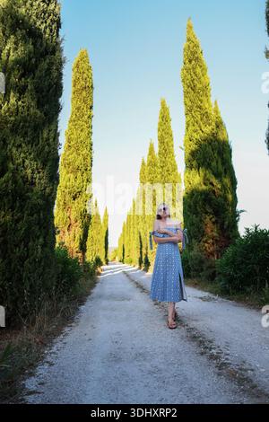 Jeune femme appréciant la promenade tranquille sur le chemin de campagne avec des arbres de cyprès de bien-être en plein air Slow Living et voyage inspiration Banque D'Images