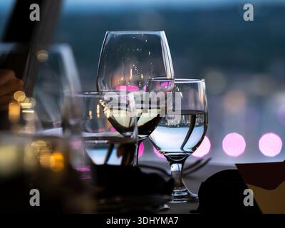Des verres à vin blanc et à eau sont assis sur une table de restaurant, reflétant la lumière ambiante et les couleurs douces de la ville. Bokeh rose et violet en arrière-plan suggèrent Banque D'Images