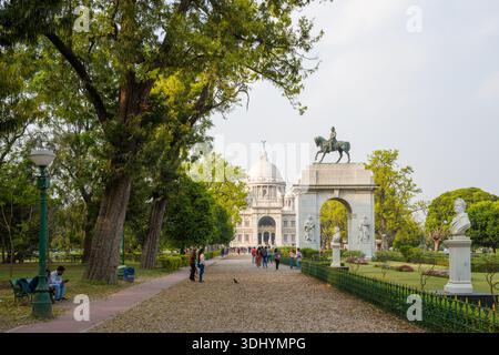 Calcutta, Inde - 23 mars 2024 : L'image montre un chemin de gravier bordé d'arbres matures et de lampadaires menant à une statue équestre sur une arche et au Mémorial Victoria, avec des gens marchant et assis sur des bancs dans les jardins environnants. Banque D'Images