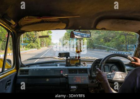 Calcutta, Inde - 24 mars 2024 : L'image montre l'intérieur d'un taxi Ambassador avec la main du conducteur sur le volant, les détails du tableau de bord et une vue à travers le pare-brise d'une large route urbaine bordée d'arbres à Calcutta. Banque D'Images