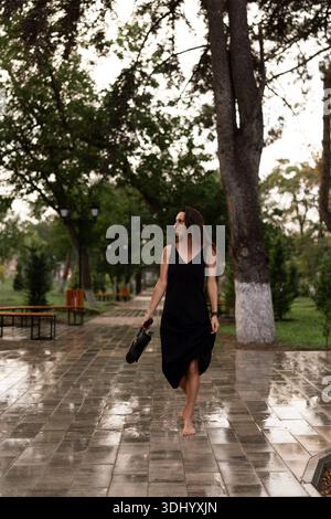 Femme, pieds nus, pluie, marche joyeusement tenant des chaussures sur un chemin de parc humide pendant une douce douche. Banque D'Images