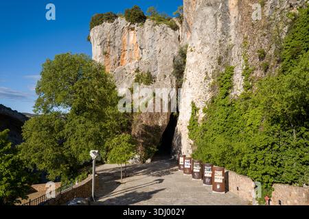 Alquezar, Espagne - 6 mai 2024 : vue sur un sentier pavé menant à l'entrée d'une grotte à la base de hautes falaises de calcaire, avec des panneaux d'affichage informatifs et la verdure environnante sous la lumière du jour clair. Banque D'Images