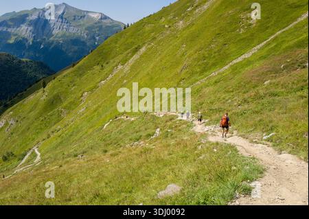 Chemin de randonnee aux chalets du Miage, France - 24 août 2024 : plusieurs randonneurs sont visibles marchant le long d'un sentier de terre sinueux sur un flanc de montagne herbeux escarpé avec un pic rocheux et une vallée boisée en arrière-plan sous un ciel bleu clair. Banque D'Images