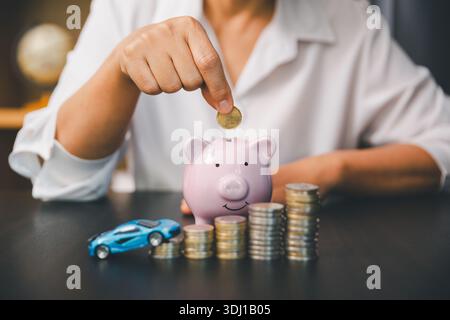 Main mettant la pièce dans la tirelire rose à côté de la voiture jouet et pièces empilées, montrant les économies de voiture, les objectifs de versement initial, le budget de prêt auto et la personne intelligente Banque D'Images