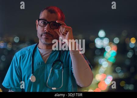 Médecin masculin en gommages ajustant les lunettes, avec des lumières de la ville en arrière-plan bokeh Banque D'Images