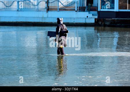 Un plongeur professionnel portant une combinaison noire et un équipement de plongée complet, y compris des réservoirs d'oxygène et des palmes, patère soigneusement dans l'eau calme et peu profonde. Banque D'Images