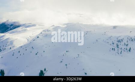 Vue aérienne large des pistes de montagne enneigées avec remontées mécaniques, lumière douce de l'hiver et nuages atmosphériques, idéal pour le tourisme d'hiver et la station alpine Banque D'Images