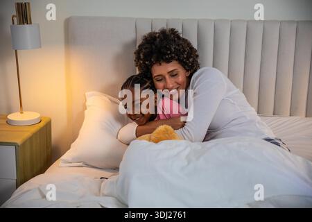 Mère et fille allongées ensemble dans la chambre sur le lit serrant sous couette blanche près de la lampe de chevet. Famille, intimité, chaleur, confort, coucher, affection, soins || autorisation du modèle Banque D'Images