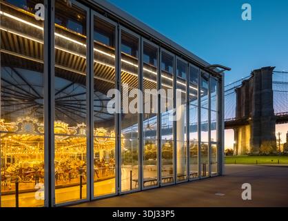 Jane’s Carousel brille au crépuscule à l’intérieur de son pavillon de verre à DUMBO, Brooklyn, avec le pont de Brooklyn s’élevant à côté de l’East River. Banque D'Images