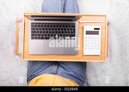 Ordinateur portable et calculatrice sur plateau dans les mains de la fille assis sur une couverture blanche douce. Flatlay, concept freelance, lieu de repos et de travail Banque D'Images