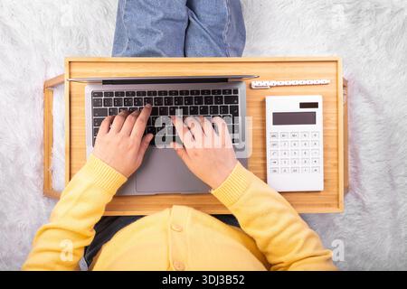 Ordinateur portable et calculatrice sur plateau dans les mains de la fille assis sur une couverture blanche douce. Flatlay, concept freelance, lieu de repos et de travail Banque D'Images