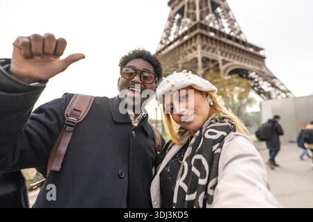 Heureux couple diversifié souriant dans la caméra tout en prenant un selfie devant la tour eiffel, profitant des voyages, du romantisme et du tourisme à paris Banque D'Images