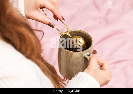 Préparation de thé vert avec infuseur doré. Mains de femme tenant la tasse et la passoire à thé. Rituel de boisson chaude maison. Moment de style de vie confortable à la maison. Banque D'Images