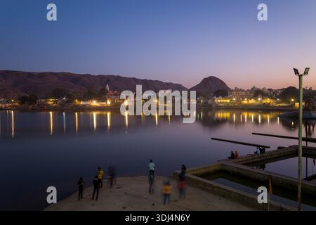 Pushkar, Inde - 18 mars 2022 : des gens sont vus debout et assis sur les ghats près du lac Pushkar au crépuscule, avec des reflets de bâtiments urbains illuminés et de collines visibles à travers l'eau calme. Banque D'Images