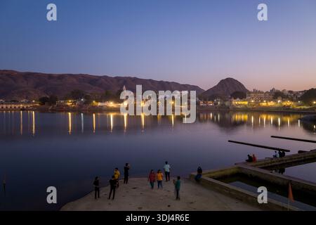 Pushkar, Inde - 18 mars 2022 : des gens sont vus debout et assis sur les ghats près du lac Pushkar au crépuscule, avec des reflets de bâtiments urbains illuminés et de collines visibles à travers l'eau calme. Banque D'Images