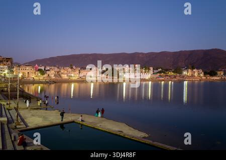 Pushkar, Inde - 18 mars 2022 : des gens sont vus debout et marchant le long des ghats près du lac Pushkar au crépuscule, avec des bâtiments illuminés et des collines reflétés dans l'eau calme. Banque D'Images