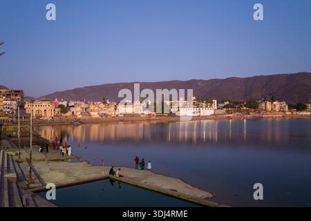Pushkar, Inde - 18 mars 2022 : les ghats et les bâtiments historiques de Pushkar se reflètent dans les eaux calmes du lac Pushkar au crépuscule, avec des gens visibles le long du bord du lac et des collines en arrière-plan. Banque D'Images